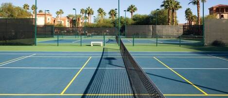 Ein Tennisplatz im Freien mit mehreren Spielfeldern, Bänken und Palmen im Hintergrund.