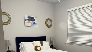 Un dormitorio ordenado con cama, ventilador de techo, ventana con persianas y una alfombra.