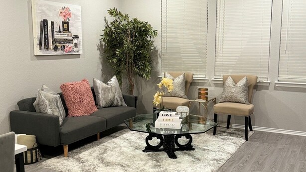Una sala de estar moderna con un sofá gris, una mesa de centro de vidrio y un sillón color beige. Hay un ventilador de techo, una planta en maceta y un cuadro en la pared.