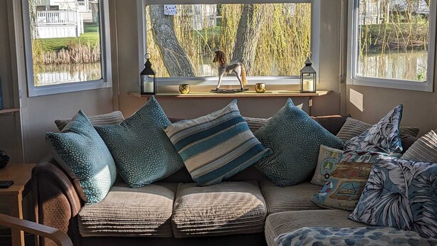 A cozy living room with a sofa set, a small table, and a window with a view of a lake and trees. - Beautiful 2 Bed Caravan Southview Park Skegness (Skegness)