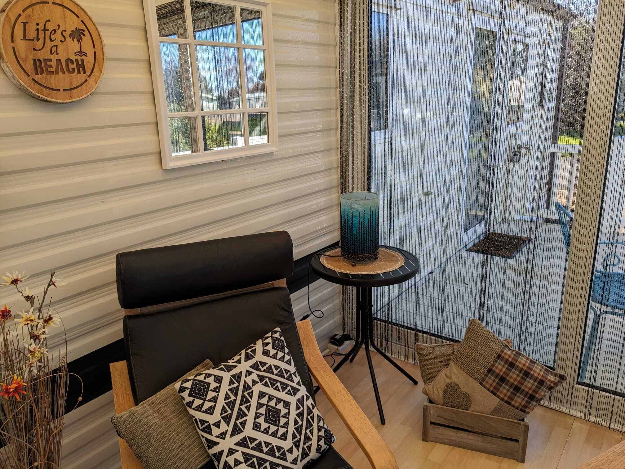 A cozy room with a black leather chair, a patterned pillow, a small round table with a candle, and a view of the outside through a window with a striped curtain.