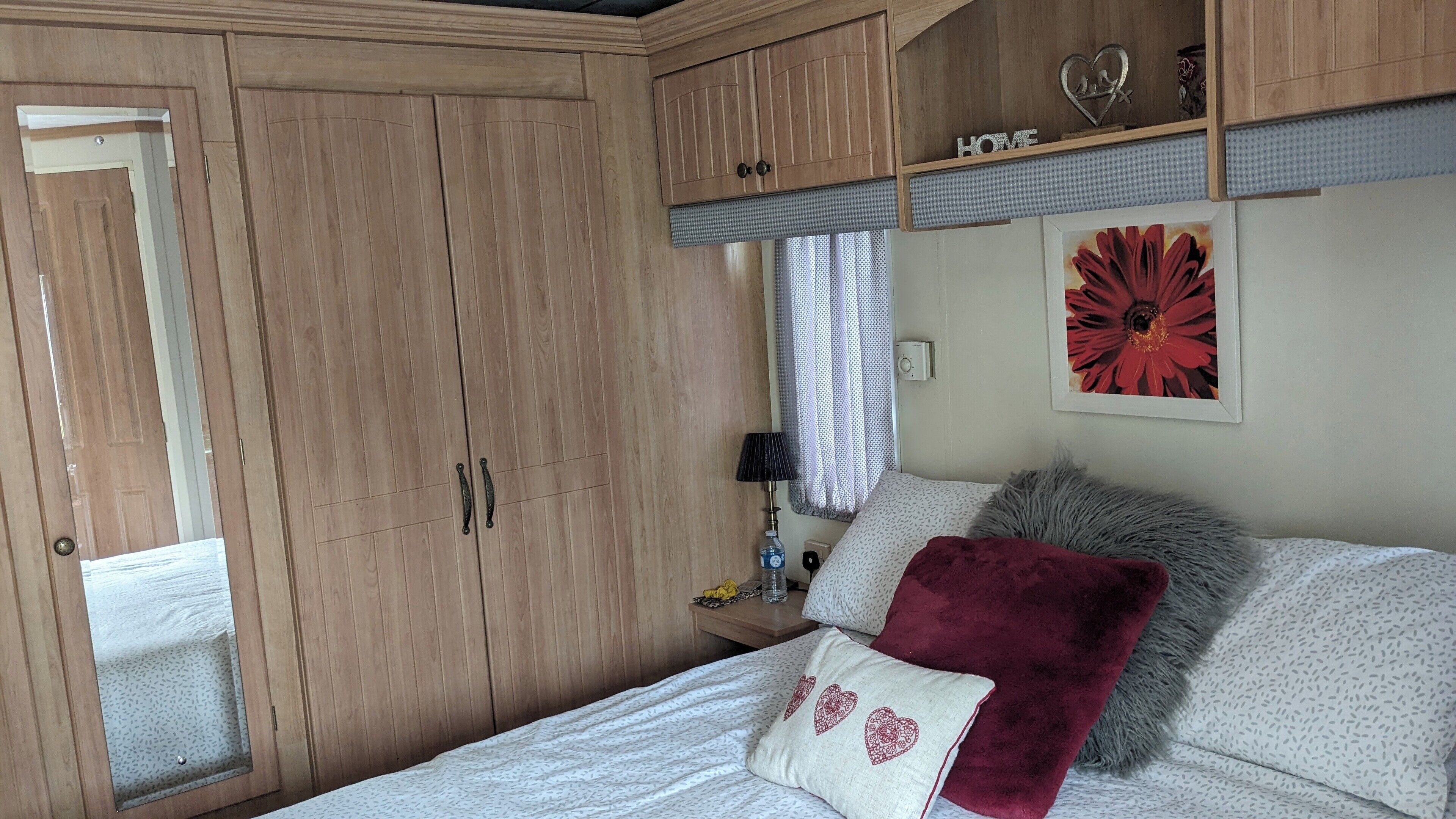 Un dormitorio con cama, almohadas, mesita de noche, lámpara, ventana con cortinas y armarios de madera.