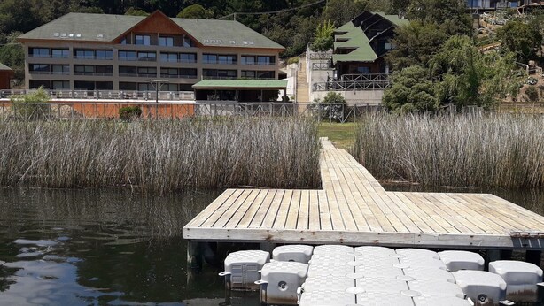 A wooden dock extending into a calm body of water, surrounded by tall reeds. In the background, there are buildings with multiple windows and a green roof, indicating a residential or hotel area. - Habitamar Lago Vichuquen (Vichuquen)