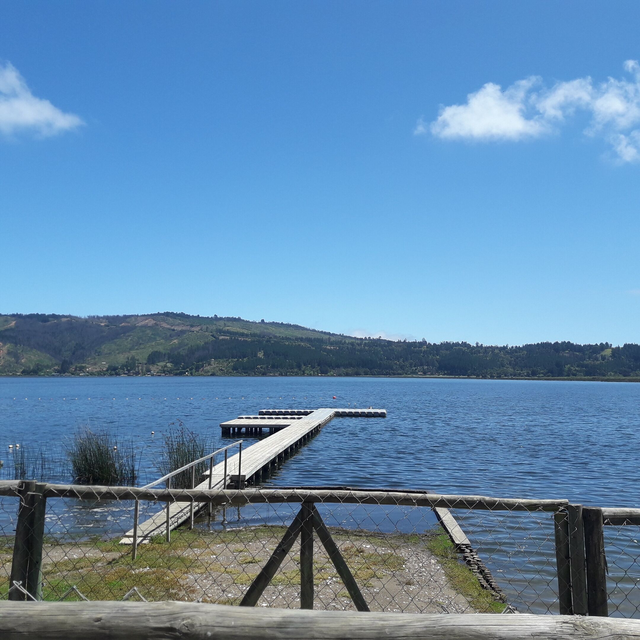 Um cais de madeira que se estende por um lago calmo, com céu limpo e colinas ao fundo.