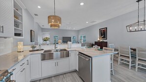Una cocina moderna con armarios blancos, una isla central, electrodomésticos de acero inoxidable e iluminación colgante.