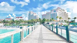 A wooden boardwalk leading to a beach with clear turquoise water, lined with palm trees and overlooking a row of high-rise hotels with red roofs. - COMPLIMENTARY CAR! CASA DEL MAR! (Nassau)