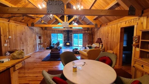 A cozy cabin interior with wooden walls and a high ceiling. There is a fireplace in the background, a round table with chairs, and a bookshelf.