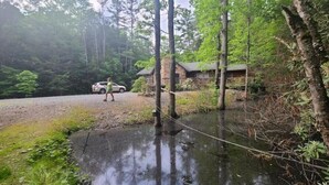 A rustic cabin with a gravel driveway and a wooden bridge over a small stream.