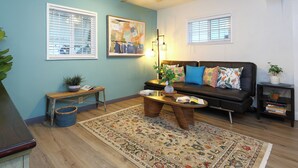 A living room with a sofa, coffee table, and a large rug. There are two windows with blinds, a ceiling fan, and a wall-mounted artwork. - Stylish 2B2B Hideaway 10 mins. drive to Disneyland (Santa Ana)