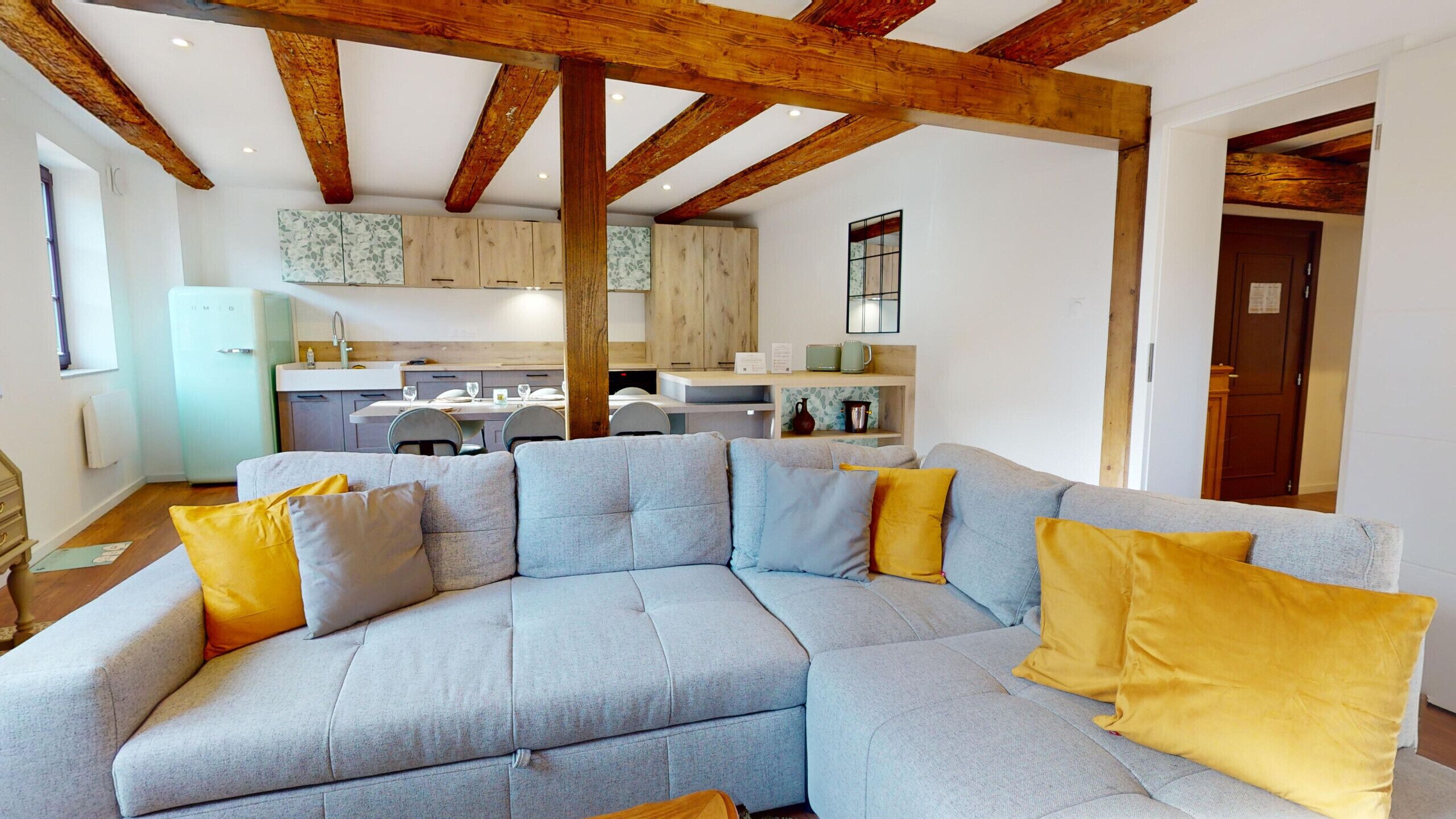 A modern living room with a grey sectional sofa, wooden beams on the ceiling, a kitchen in the background, and a door to the right.