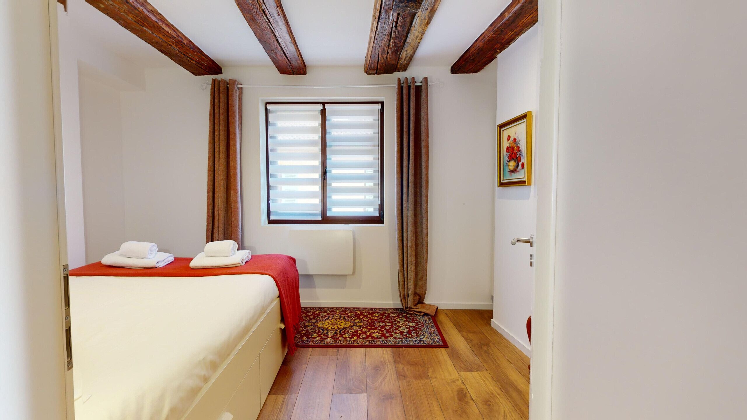 Een hotelkamer met een bed, een raam met zonwering, een deur en een ingelijst kunstwerk aan de muur.