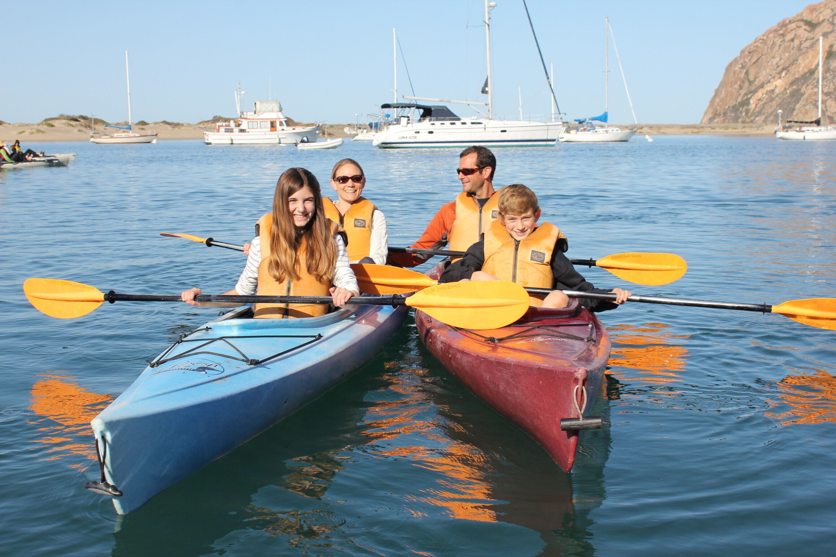 A group of people in kayaks on calm water with boats in the background.