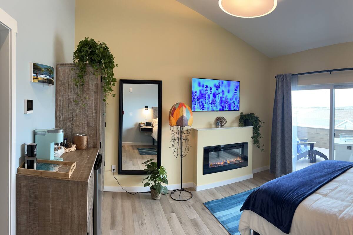 Un dormitorio moderno con cama, chimenea, un ventanal con cortinas, televisor y espejo.