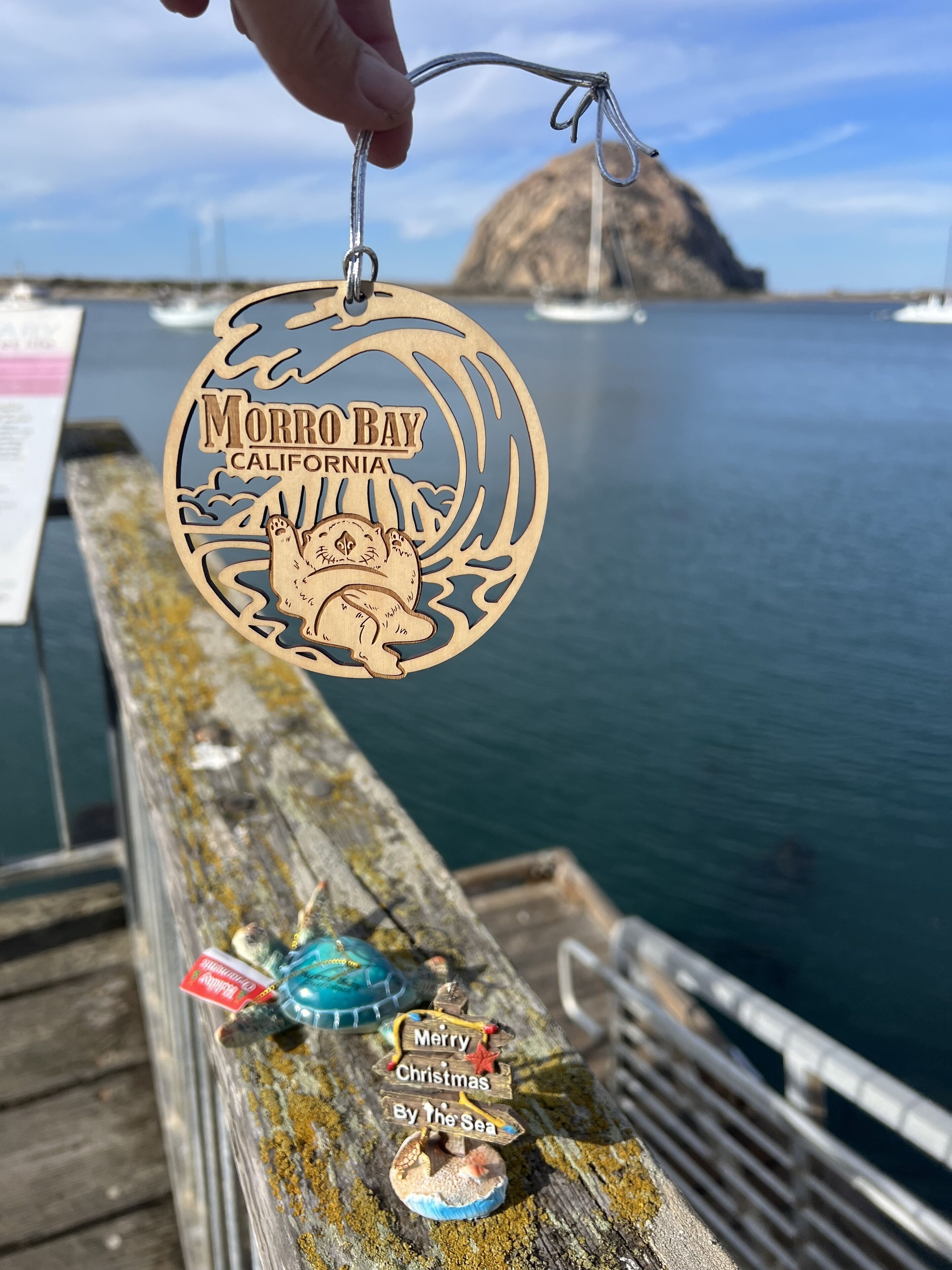 A wooden keychain with "Morro Bay California" text and a bear design, held over a wooden railing with a view of the sea and a sign that reads "Merry Christmas By The Sea".
