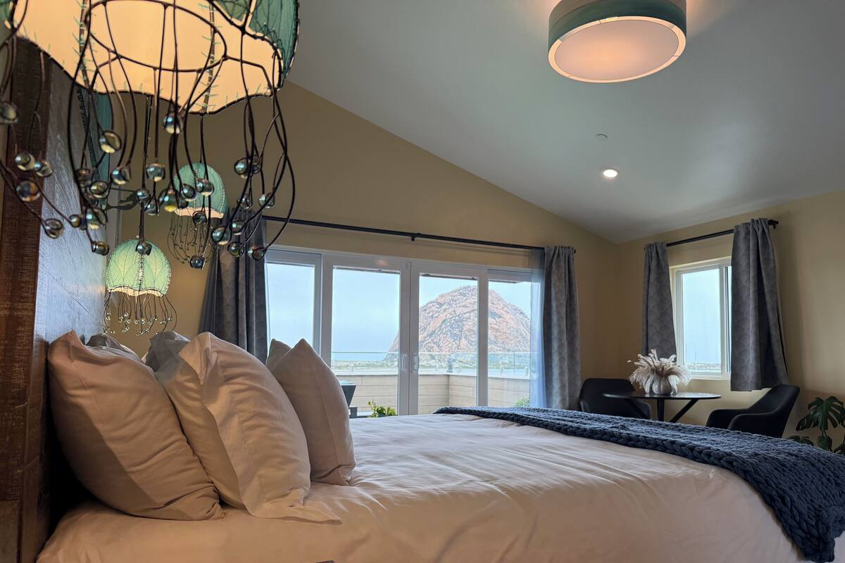 Habitación de hotel con una cama grande, un escritorio con silla y vistas al océano a través de las ventanas.