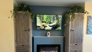 A living room with a fireplace, a television mounted above it, and wooden cabinets with plants on top. - HarborWalk Inn (Morro Bay)