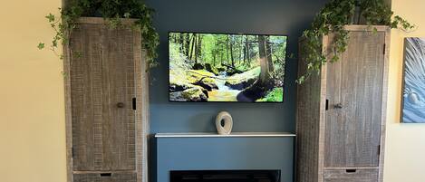 A living room with a fireplace, a television mounted above it, and wooden cabinets with plants on top.
