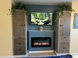 Una sala de estar con chimenea, televisor montado encima y armarios de madera con plantas arriba.