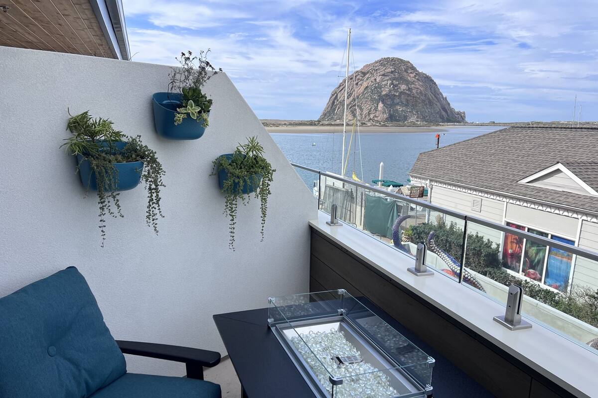 Un balcón con vista al mar, una silla, una mesa con vitrina de vidrio y plantas colgantes.