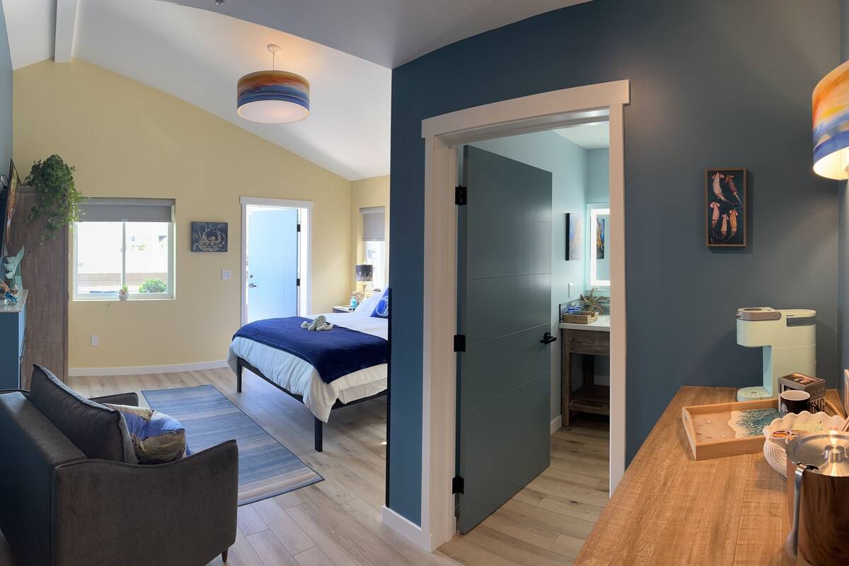 Un dormitorio moderno con una cama grande, una zona de estar con un sofá y un baño visible a través de una puerta abierta.