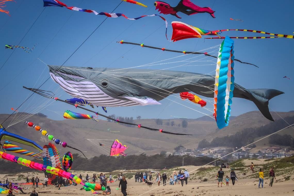Una playa con numerosos cometas volando en el cielo y gente caminando sobre la arena.