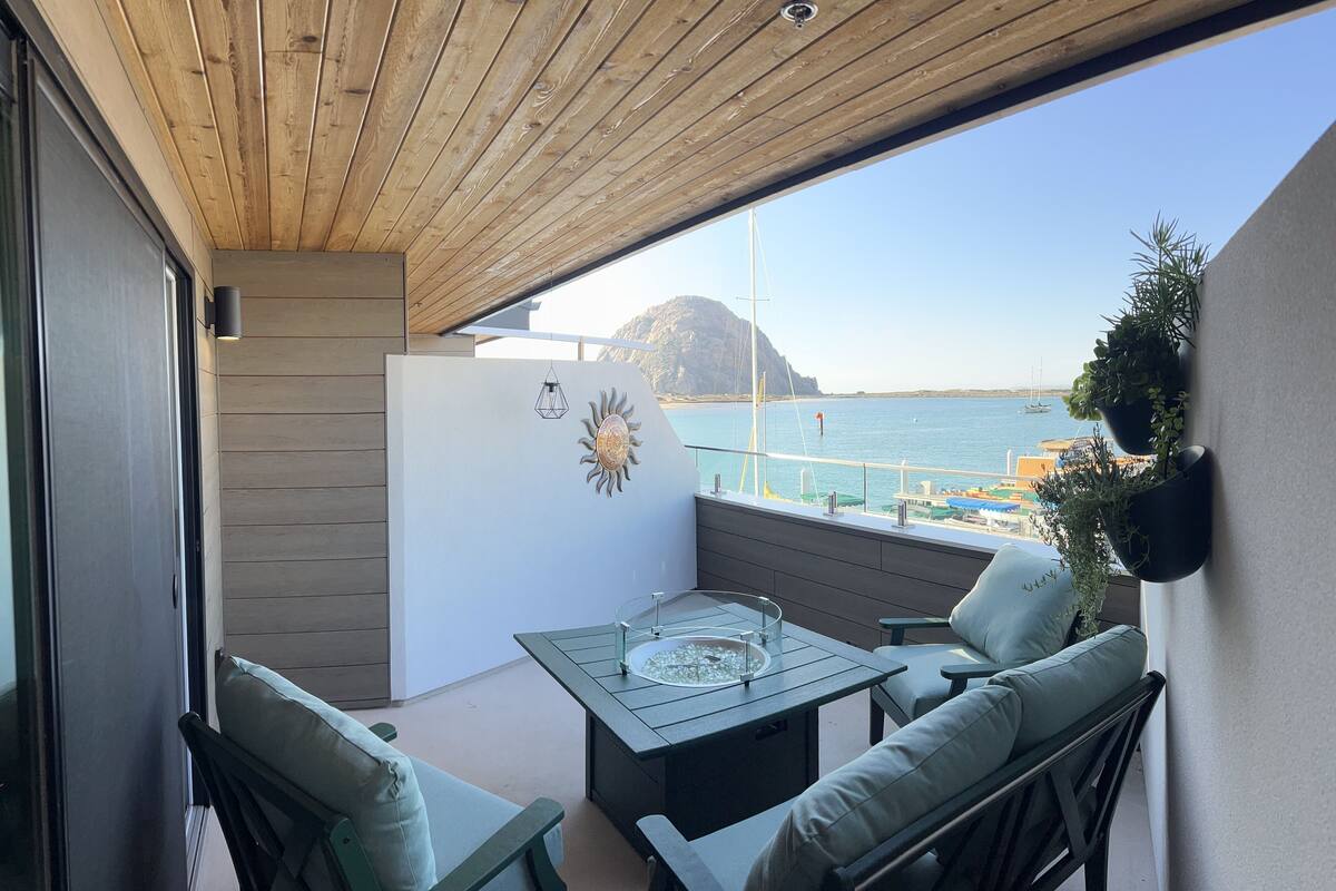 Un balcón con techo de madera, una mesa de vidrio y dos sillas con cojines verdes. Se observa un cuadro decorativo de un sol, y el balcón da a una zona costera con barcos y edificios.
