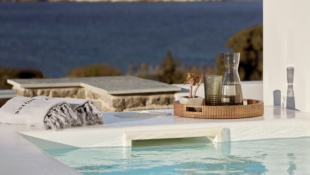 A pool area with a view of the sea and mountains, a towel, and a tray with a vase and a candle. - Manesis Suites (Milos)