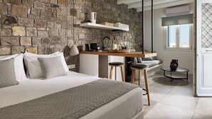 A bedroom with a stone wall, a bed with patterned bedding, a wooden desk with stools, and a window with a view. - Manesis Suites (Milos)