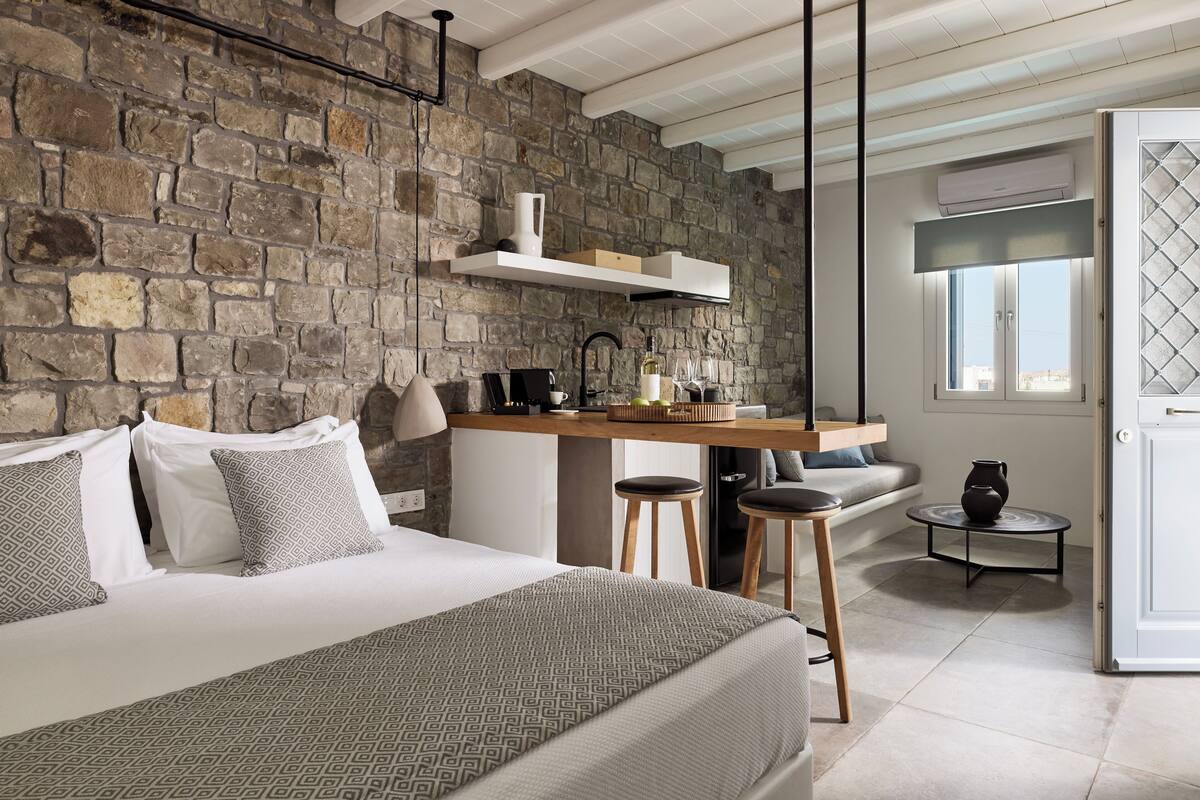 A bedroom with a stone wall, a bed with patterned bedding, a wooden desk with stools, and a window with a view.