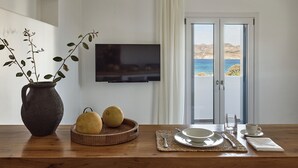 A dining area with a wooden table, a vase with plants, a fruit bowl, a TV mounted on the wall, and a view of the sea through a glass door. - Manesis Suites (Milos)