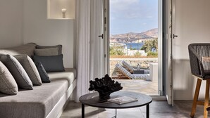 A modern living room with a sofa, a round table with a vase, and a view of a balcony with outdoor furniture. - Manesis Suites (Milos)