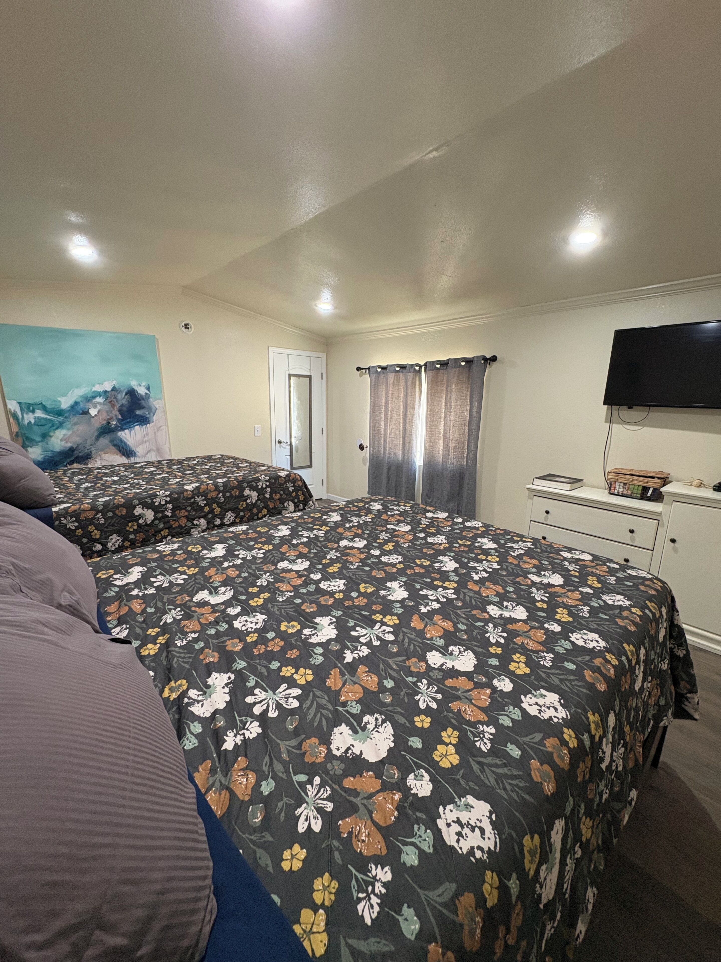 A hotel room with two beds, a flat-screen TV, a painting on the wall, and a door with curtains.