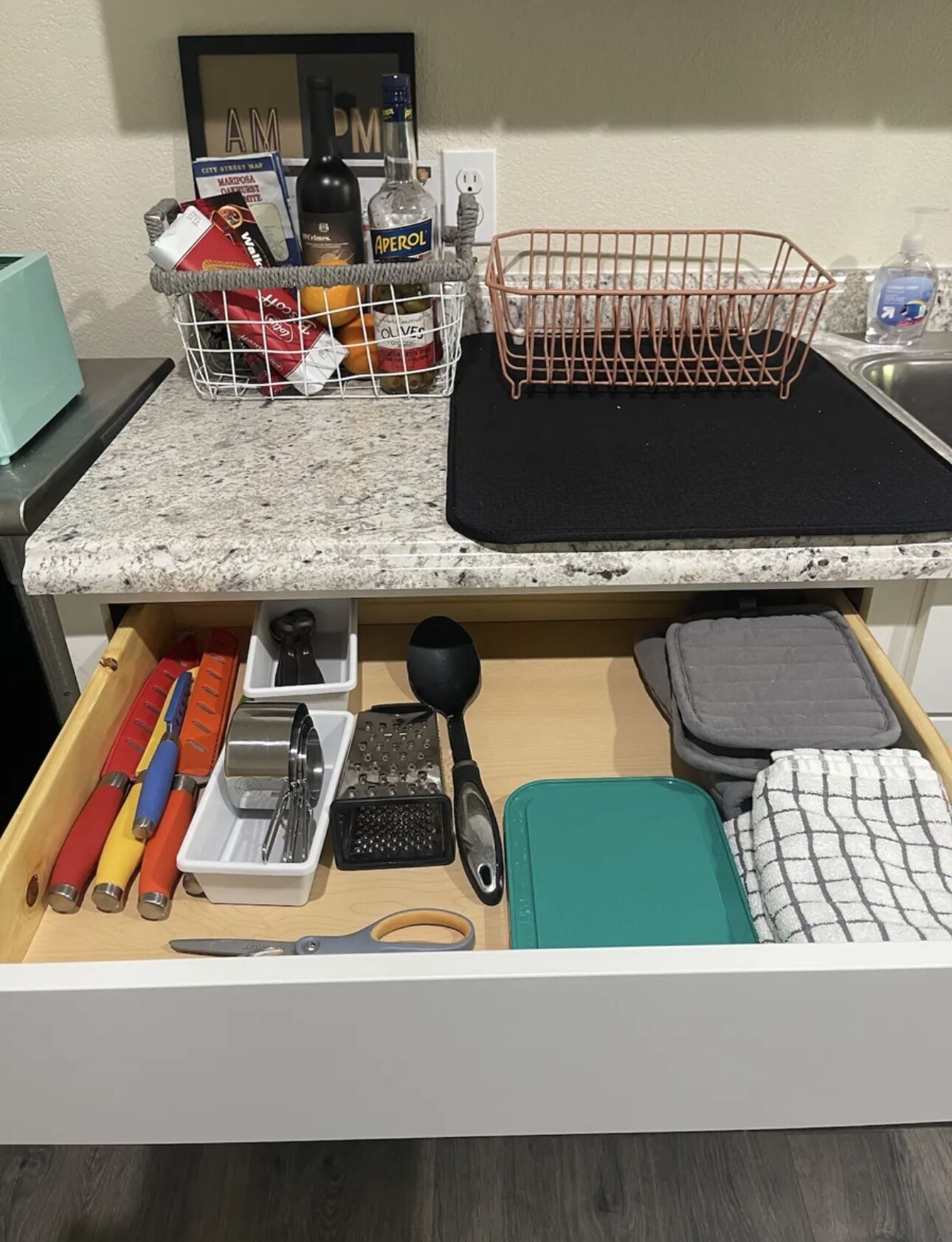 A kitchen counter with a marble top, a wire basket, a black mat, and a drawer with kitchen utensils.