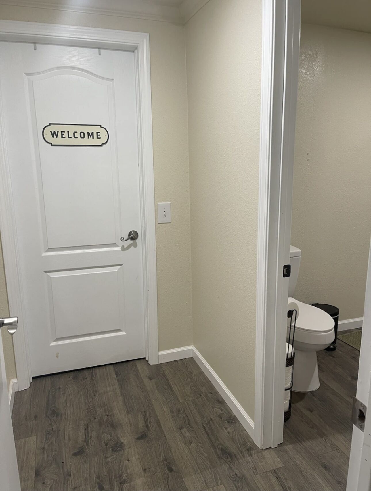 Een witte deur met een “WELCOME” bord, een houten vloer en een wit toilet in een kleine badkamer.