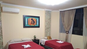 A hotel room with two beds, each with a red bedspread and a towel folded on top. There is a painting on the wall, a small table with a plant, and a window with curtains. - Hotel Vanderkris Chone (Chone)