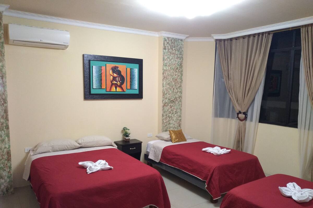 A hotel room with two beds, each with a red bedspread and a towel folded on top. There is a painting on the wall, a small table with a plant, and a window with curtains.