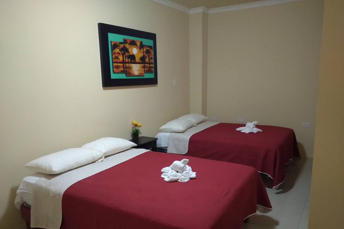 A hotel room with two beds, each with red bedding and white pillows. There is a framed picture on the wall above the beds.
