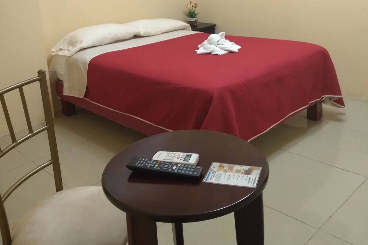 A hotel room with a bed, a small table with a remote control and a menu, a chair, an air conditioner, and a painting on the wall.