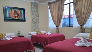 A hotel room with three beds, each with a red bedspread and white pillows. There is a painting on the wall, a window with curtains, and a view of a building outside. - Hotel Vanderkris Chone (Chone)