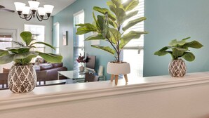Uma sala de estar com sofá, mesa de centro e lustre. Três plantas em vasos decoram uma lareira.