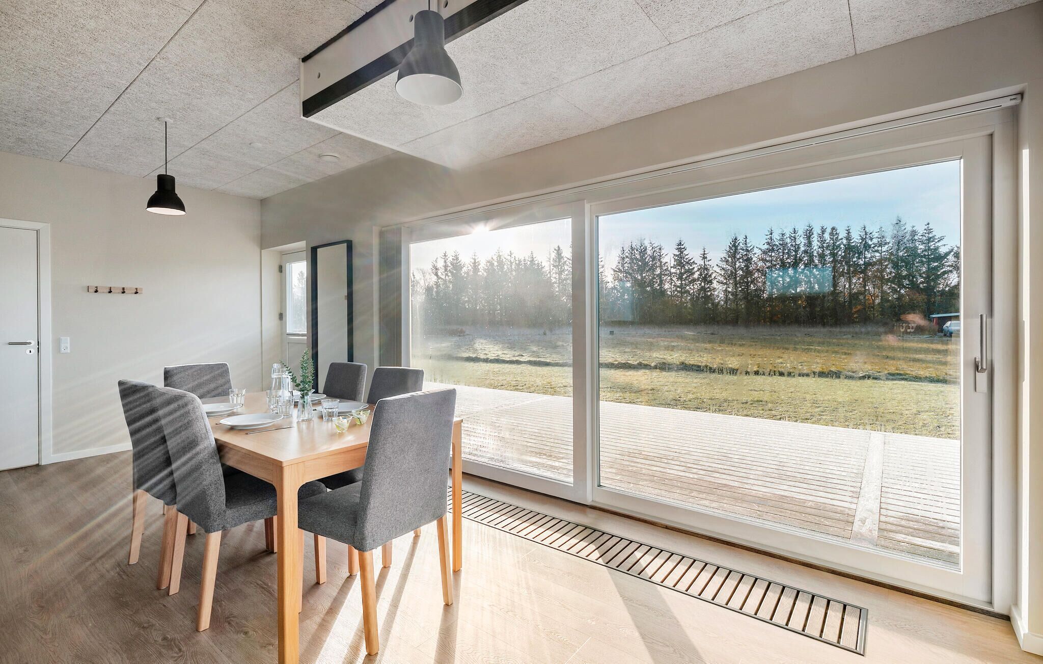 A modern dining area with a large table, grey upholstered chairs, and a view of a forest through large sliding glass doors.