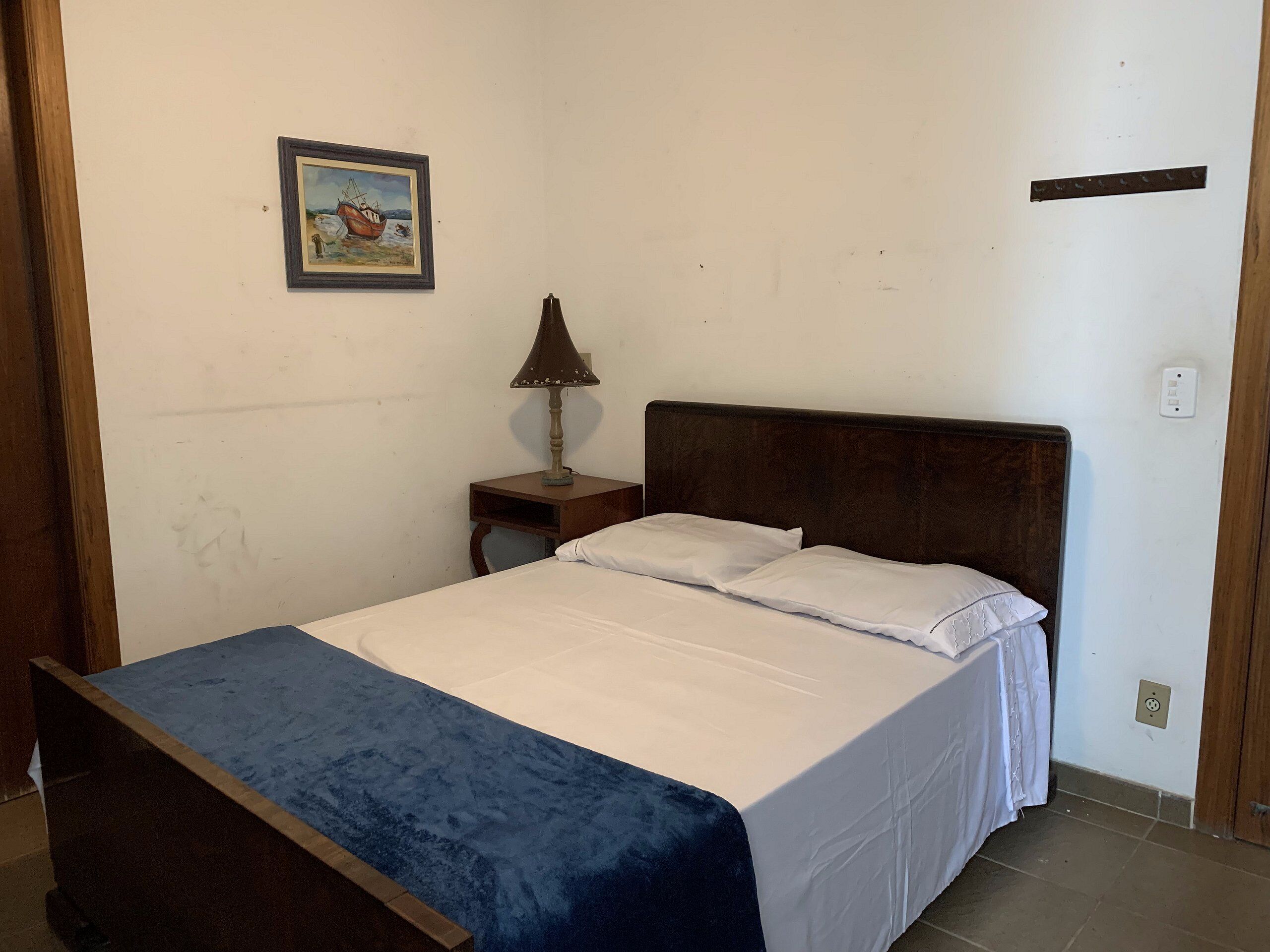 Un dormitorio con una cama de madera, ropa de cama blanca, una mesita de noche pequeña con una lámpara y un cuadro en la pared.