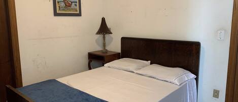 Un dormitorio con una cama de madera, ropa de cama blanca, una mesita de noche pequeña con una lámpara y un cuadro en la pared.
