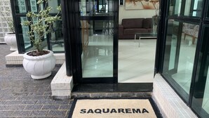 Entrada de hotel con puerta de vidrio, felpudo con la palabra "SAQUAREMA" y una planta en maceta, además de un sofá visible en el interior.