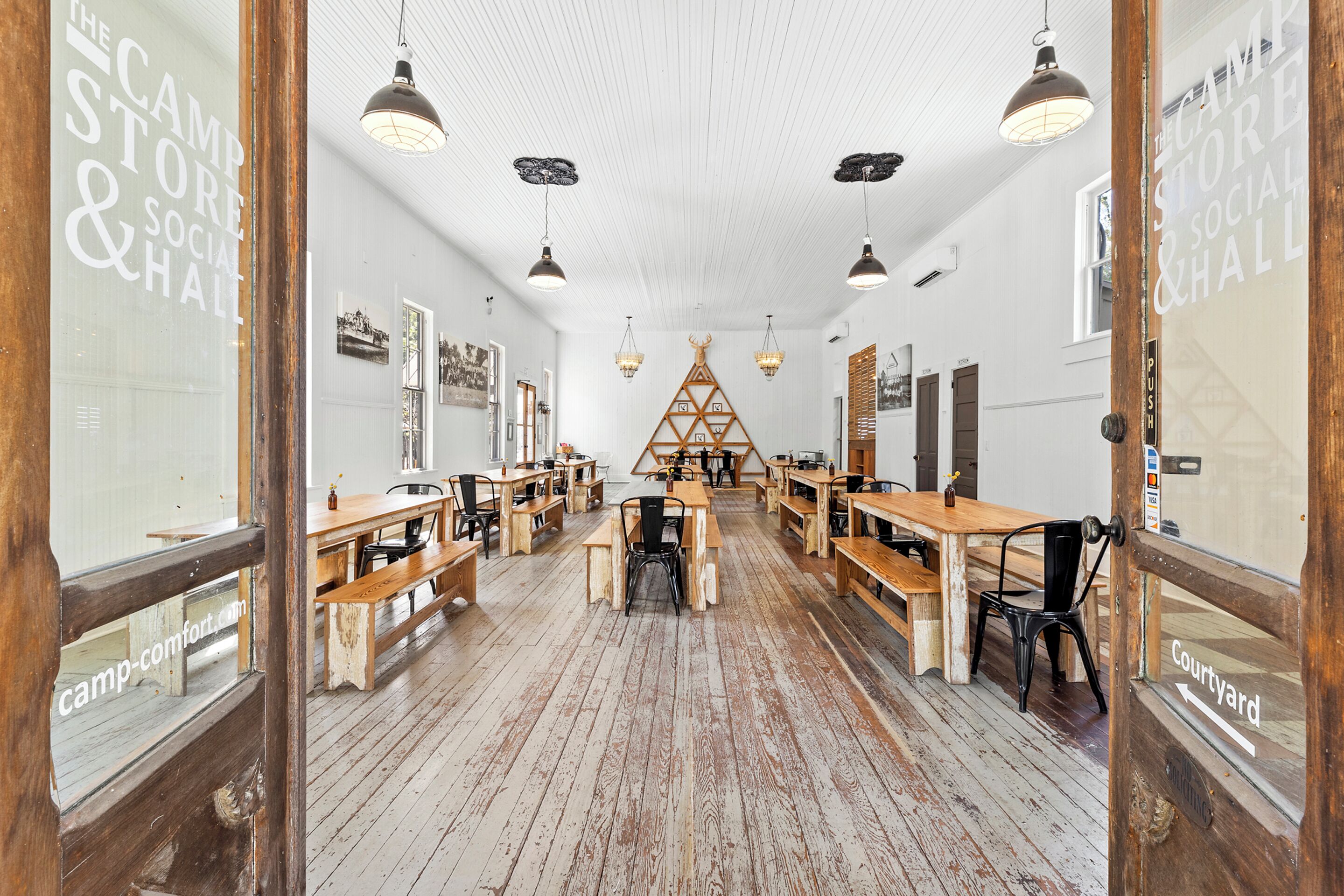 A spacious room with wooden benches and tables, white walls, and a high ceiling with pendant lighting.