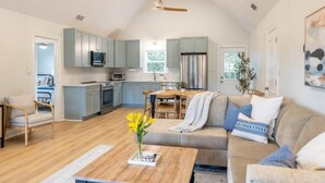 A spacious living area with a sofa, coffee table, and a dining table. There is a kitchen in the background with light blue cabinets and a ceiling fan. - Newly Renovated, 1 min walk to beach, 3rd Row Emerald Isle Beach House (Emerald Isle)