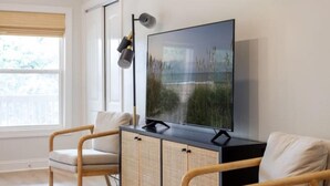 A modern living room with a large flat-screen TV mounted on a black cabinet, two wicker chairs, a floor lamp, and a window with a view of greenery. - Newly Renovated, 1 min walk to beach, 3rd Row Emerald Isle Beach House (Emerald Isle)