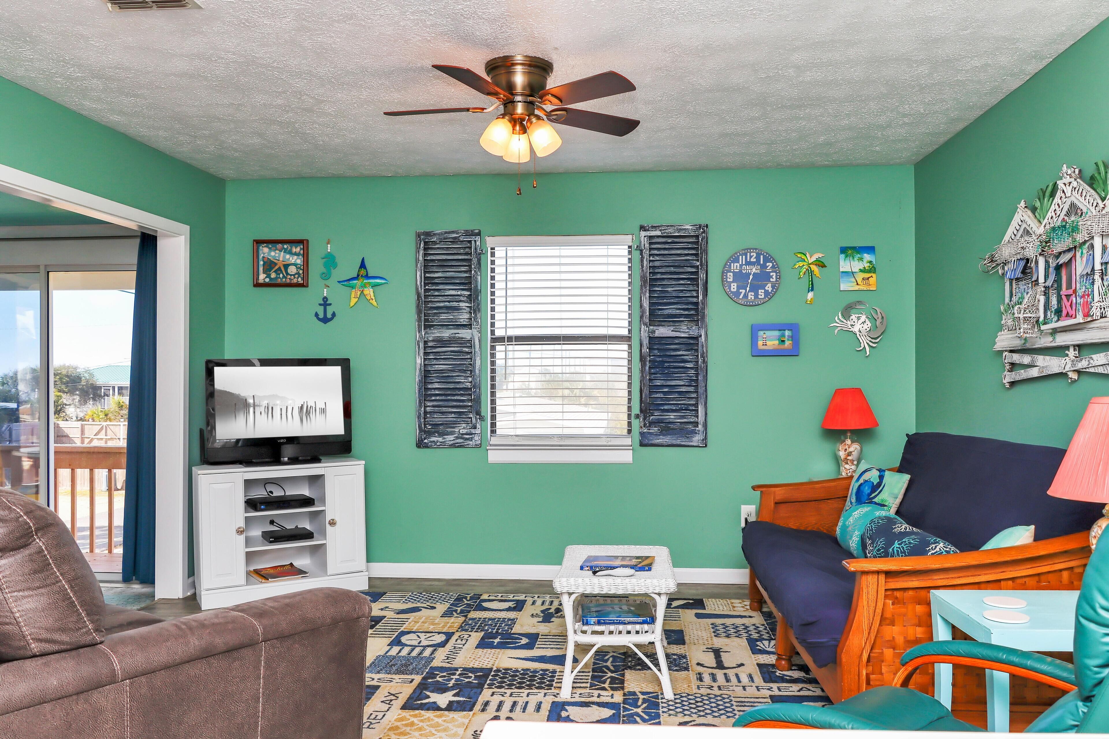 A living room with teal walls, a ceiling fan, a TV on a white stand, a wooden sofa, a small table, and a patterned rug.