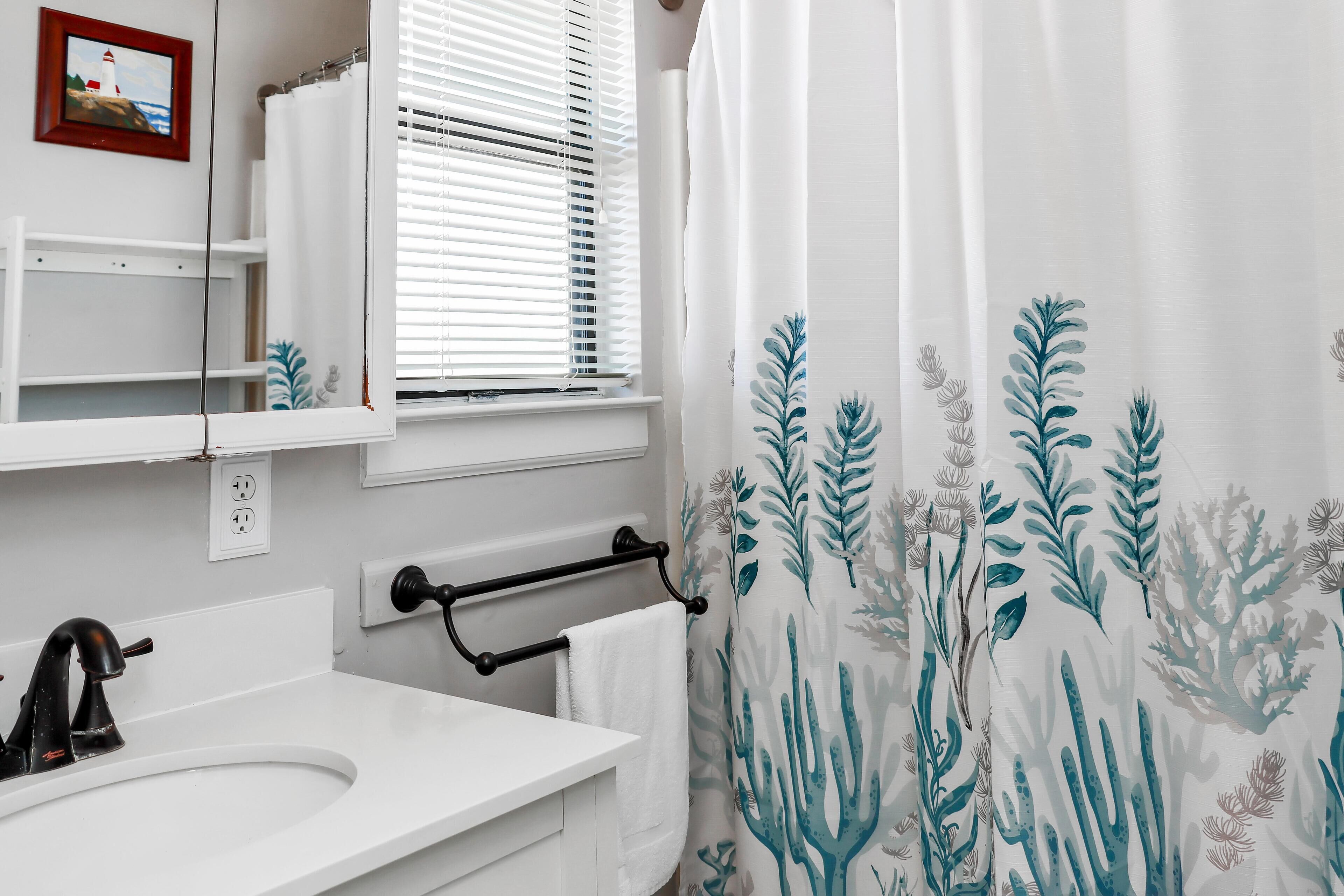 A bathroom with a white sink, black faucet, a towel rack, a window with blinds, and a shower curtain with a leaf pattern.