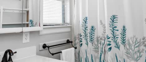 A bathroom with a white sink, black faucet, a towel rack, a window with blinds, and a shower curtain with a leaf pattern.